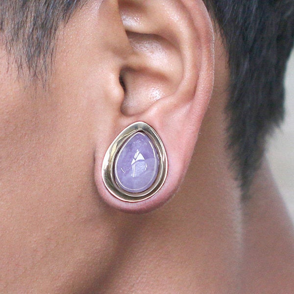 Close-up of a person's ear adorned with the Kaya Movement's Amethyst Teardrop Stone Saddle Plug in Gold. The elegant body jewelry showcases a pale lavender natural amethyst stone set in a gold bezel, perfectly complementing the person's light brown skin tone and short dark hair. This stylish gauge earring is available in sizes from 12mm to 25mm.